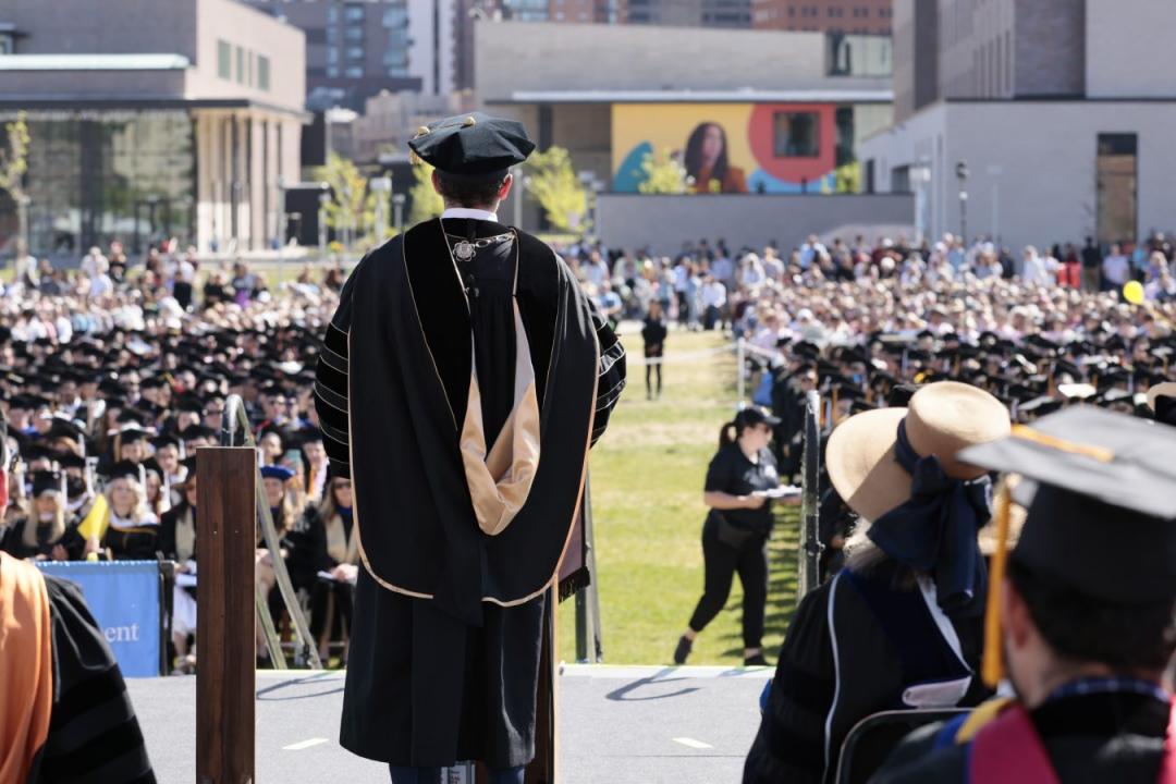 丹佛大学的户外毕业典礼照片. 照片中，一位演讲者穿着黑色毕业礼服，背对着镜头向人群演讲. 