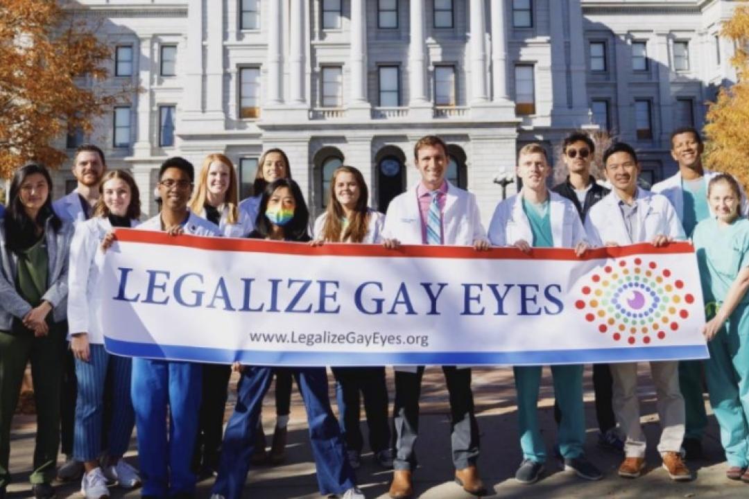 More than a dozen medical professionals stand together holding a sign that reads "Legalize Gay Eyes" in front of the Colorado State Capitol building.