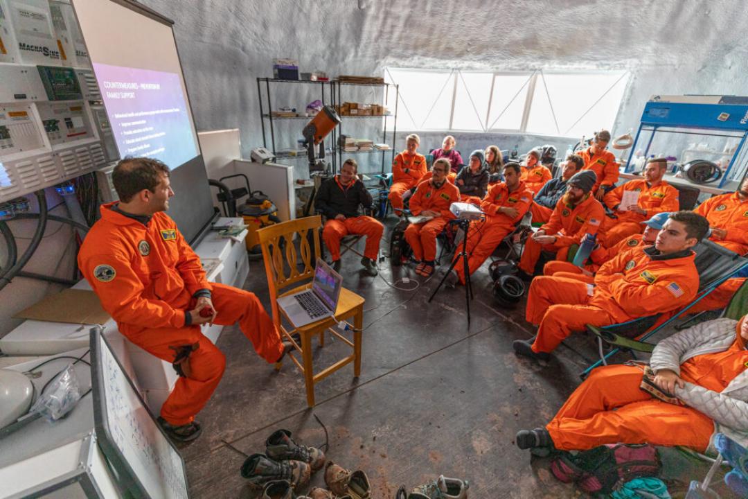Photo of one instructor and a couple dozen students, all in bright orange environment suits, gathered in a building for a presentation during a simulation exercise at the Mars Desert Research Station in 2019.