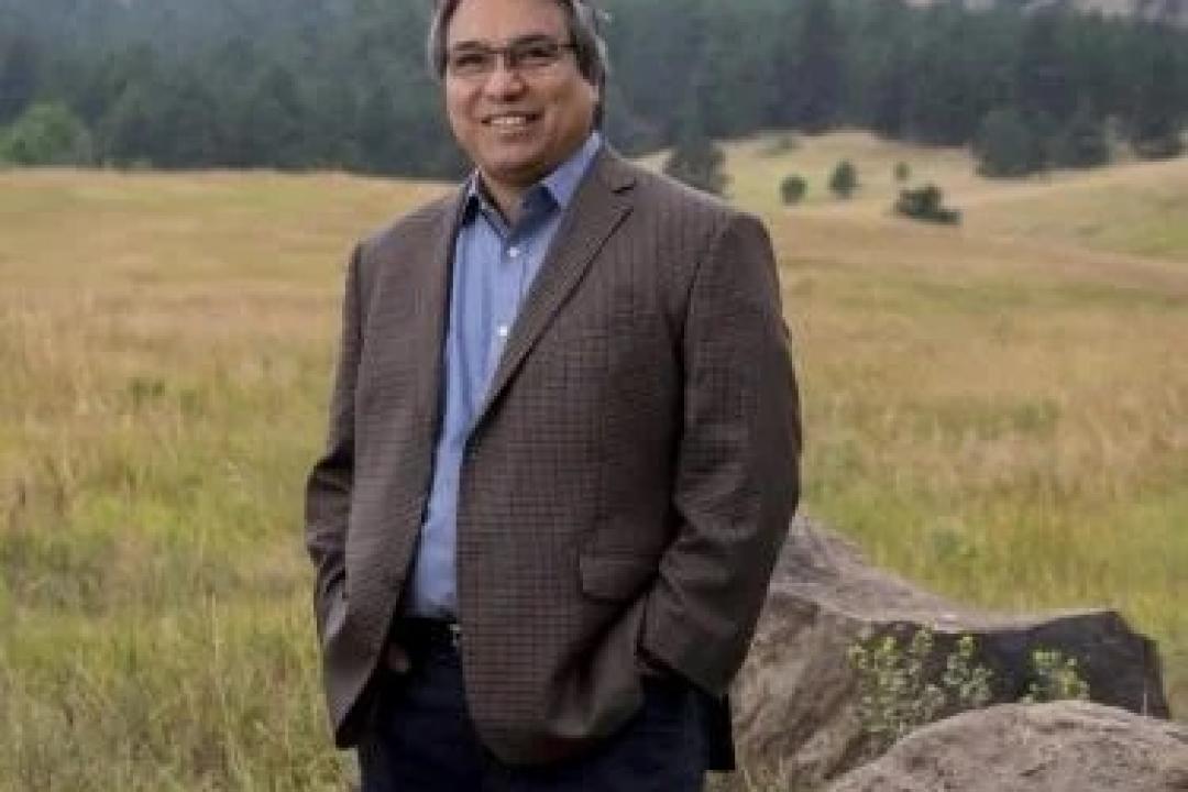 James Anaya in a brown suit jacket, blue shirt, and dark blue pants. Anaya is standing outside in a field with grass, small boulders, and mountains behind. 