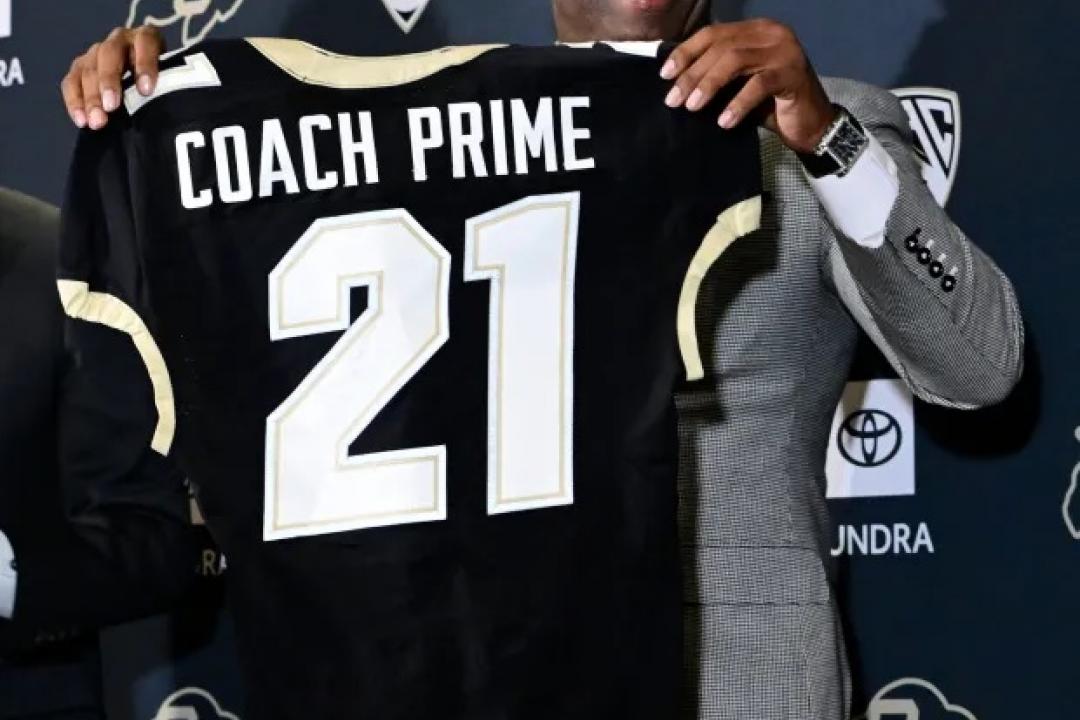 Coach Deion Sanders holding a black CU jersey with Coach Prime on the back 