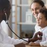 Photo of a doctor using a stethoscope to listen to the heart of a young child held on their mother's lap. 