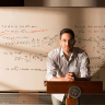 Daniel Larremore leaning over a podium with a whiteboard marked with physics notations in the back 