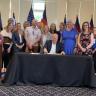 Gov. Polis at a black table signing a bill with a crowd of legislators behind him. The walls are beige and the floor is a dark blue pattern.