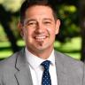 Chad Marturano in a gray suit with a blue polka dot tie. Blurred trees and greenery are in the background. 