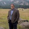 James Anaya in a brown suit jacket, blue shirt, and dark blue pants. Anaya is standing outside in a field with grass, small boulders, and mountains behind. 